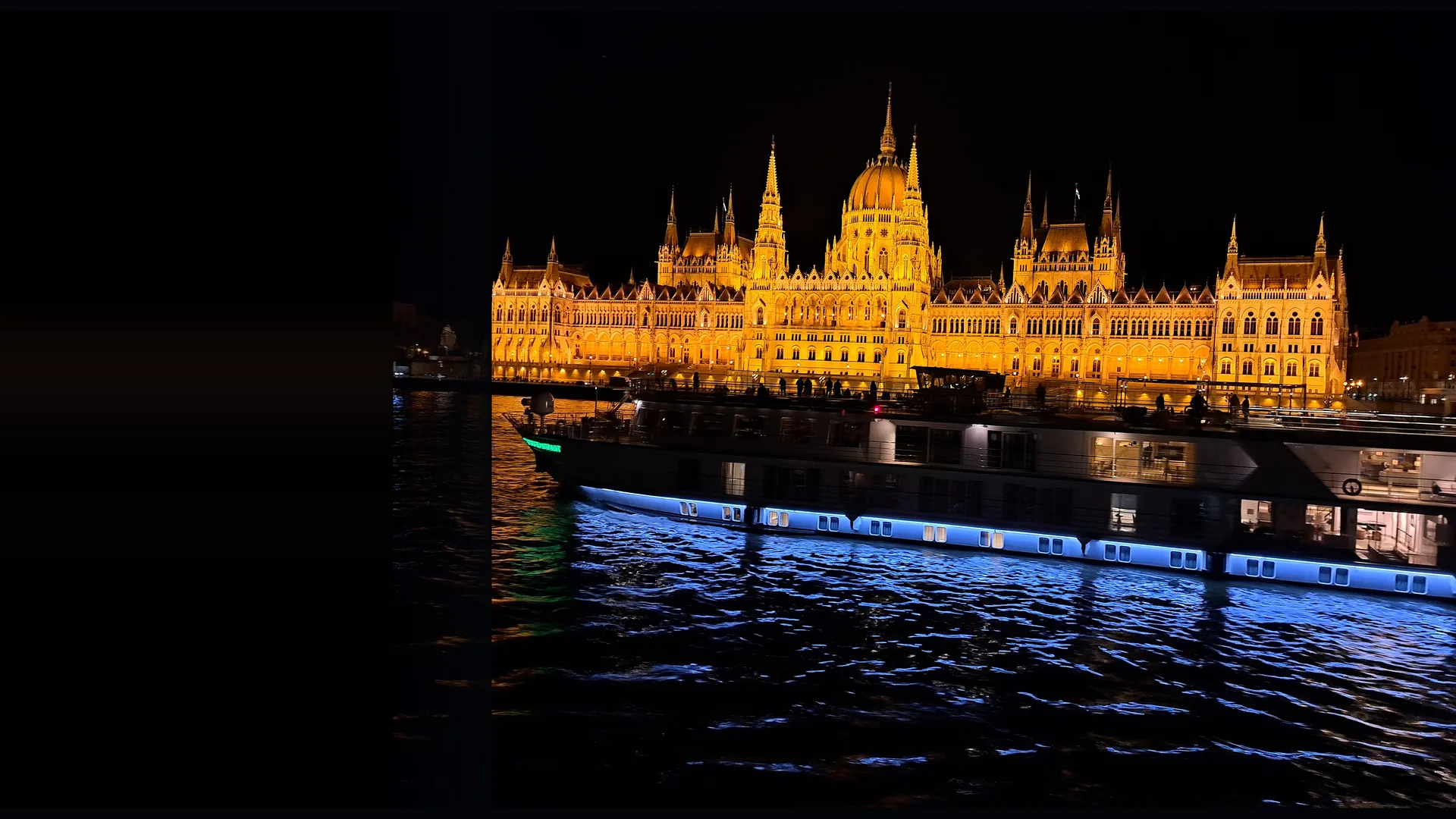 The Hungarian Parliament Building glowing gold on the Danube bank at night, with a river cruise boat outlined in blue light in the foreground.